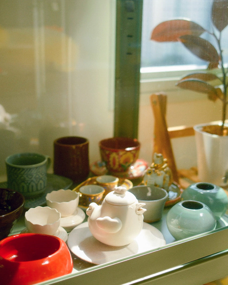 A collection of colorful ceramic tableware, including a white teapot, cups, and small vases, arranged inside a glass display cabinet with a potted plant in the background.