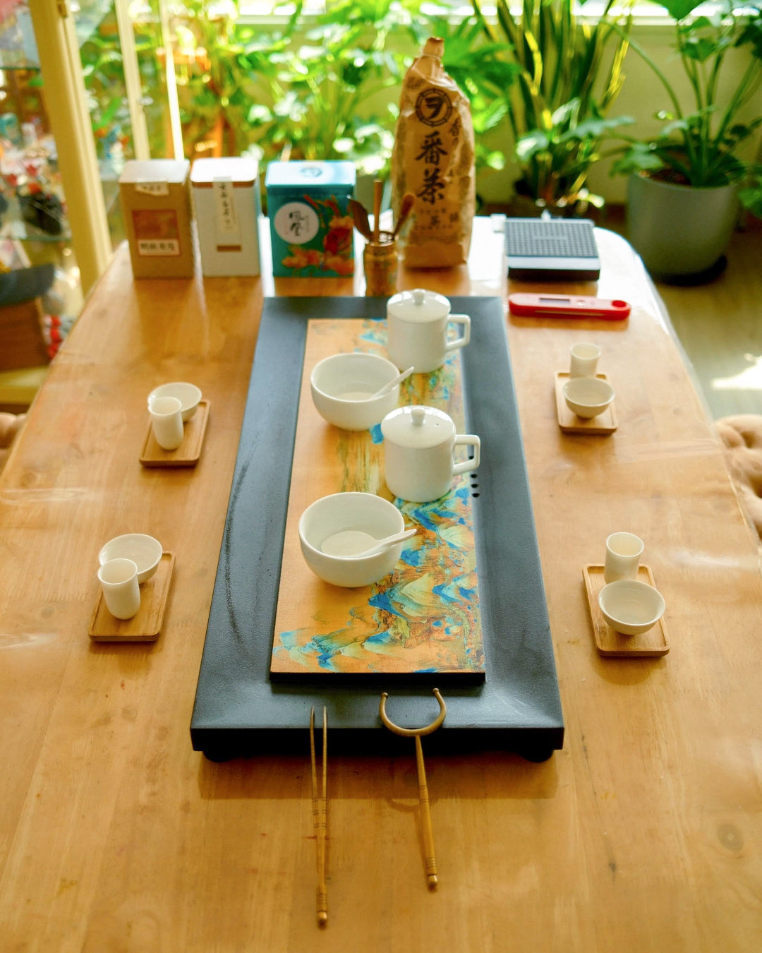 A traditional tea set arranged on a wooden table, featuring a long black rectangular tray with a colorful central panel, white ceramic teacups and bowls, small wooden saucers, and a tall paper bag of matcha leaves in the background.