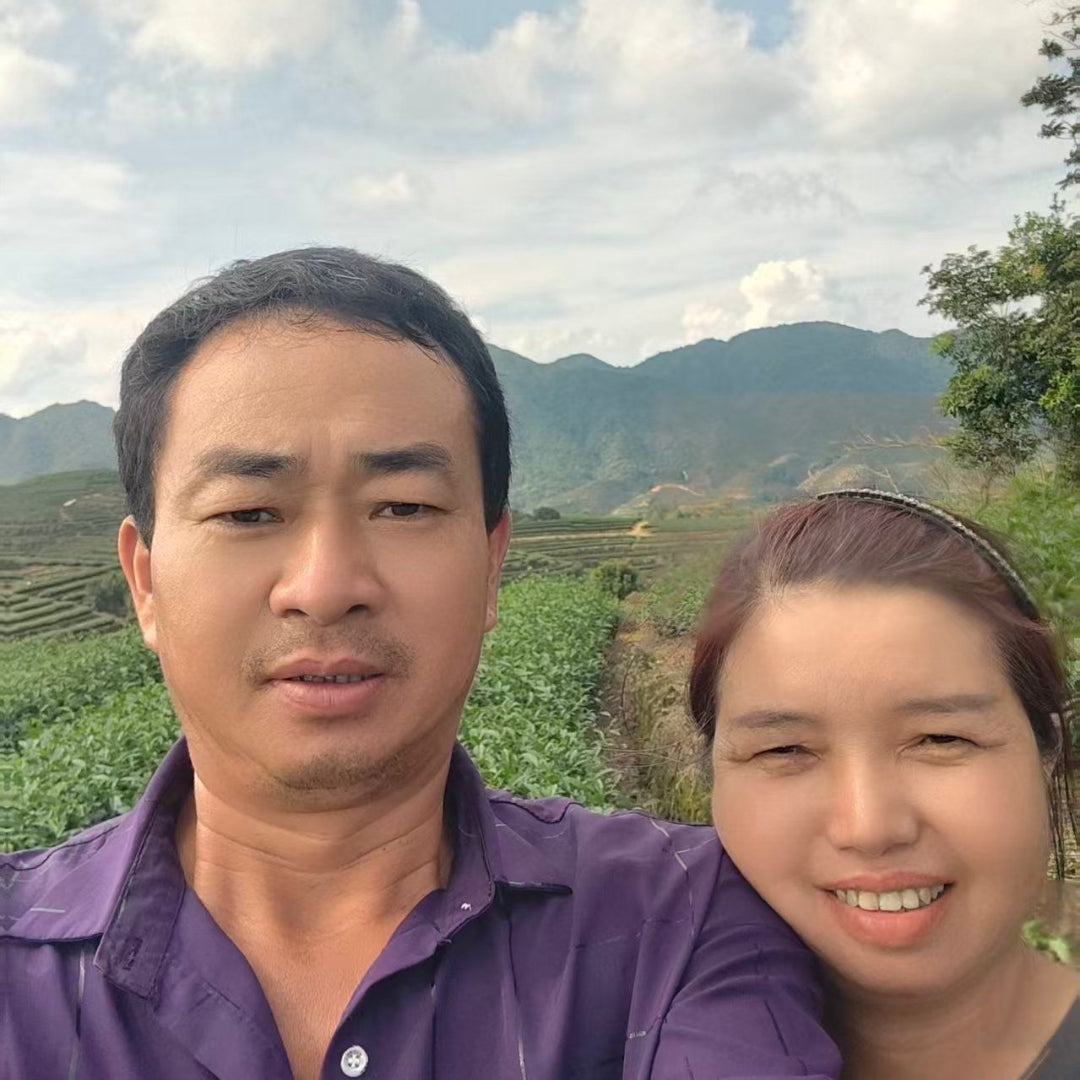 Tea master and family taking a selfie outdoors in front of mountains and tea fields under a cloudy sky in Fujian. The man is wearing a purple shirt.