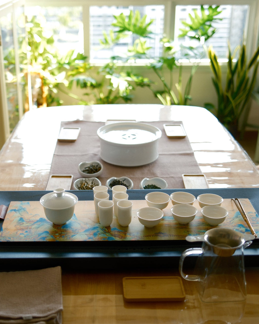 A traditional Chinese tea set arranged on a rectangular wooden table in a bright, sunlit room with potted plants in the background. The set includes a dark blue tray, white ceramic teapots and cups, small bowls, and a glass pitcher.
