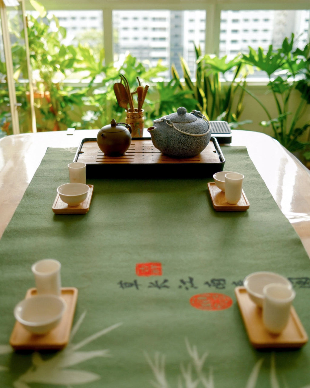 A green tea table runner with Chinese calligraphy and red seal stamps, styled with a gray teapot, bamboo utensils, and white teacups on wooden trays in front of a window with green plants.