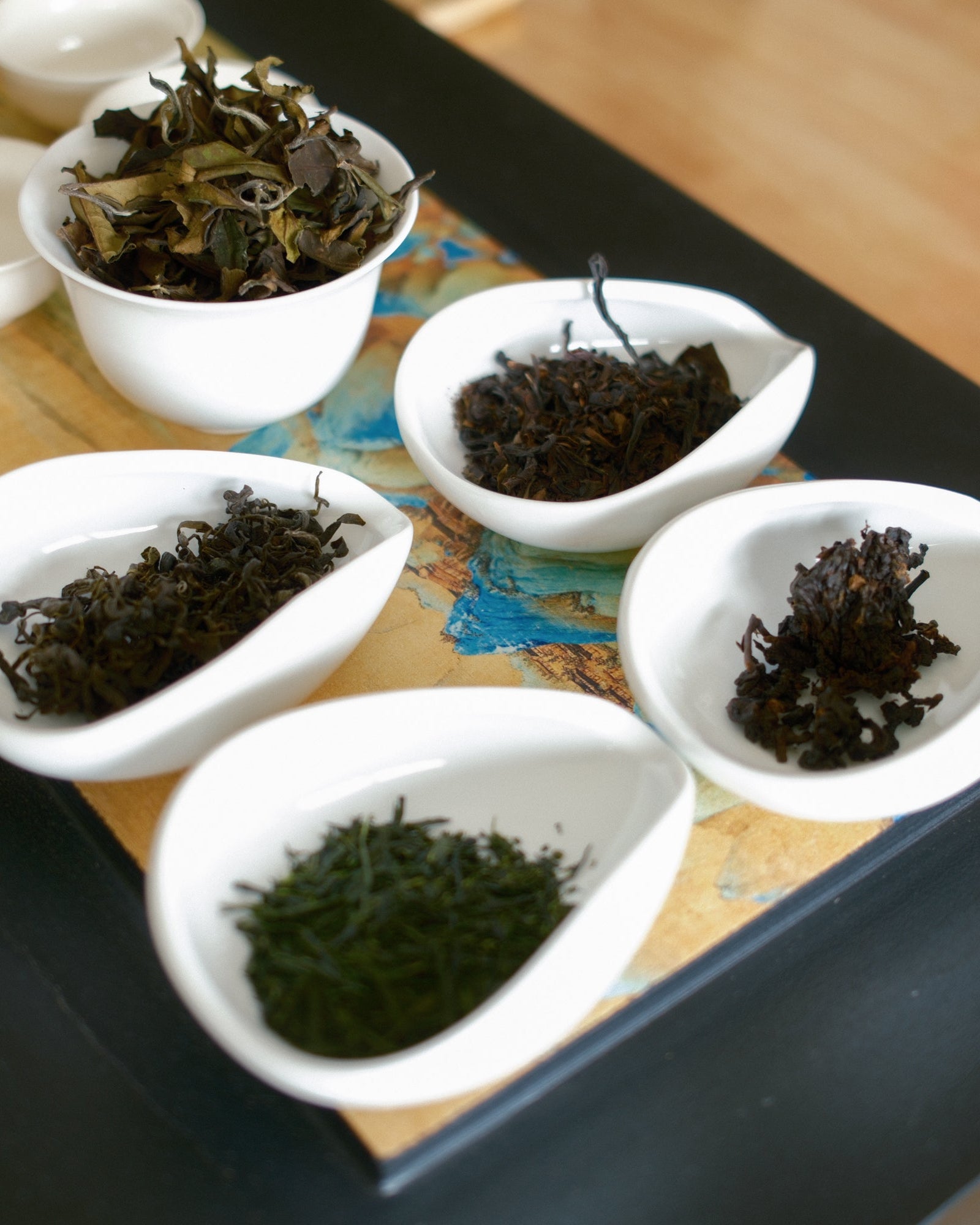 Six white ceramic teacups arranged on a black tray over a wooden surface, each holding different dried tea leaves in varying shades of green and brown.