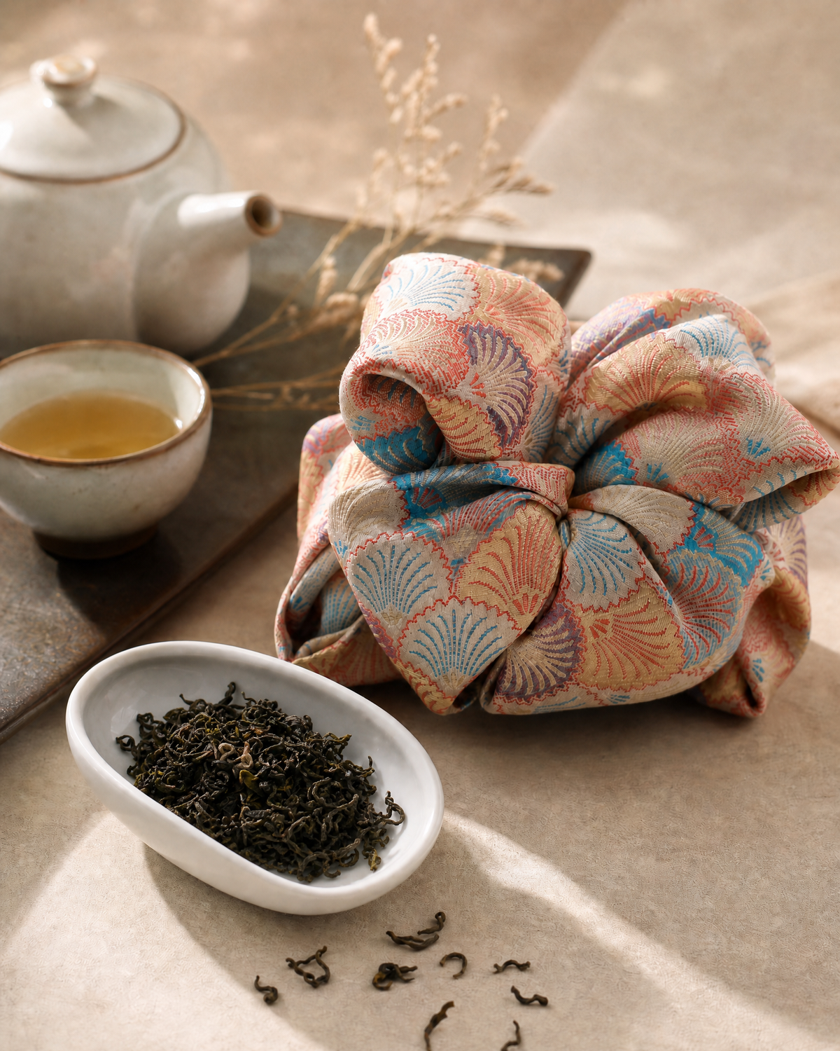 A beautifully wrapped tea gift bag from Cha Cha Chan with a pink, blue, and beige fan pattern sits on a neutral surface next to a small dish of loose-leaf tea, a cup of tea, and a teapot.