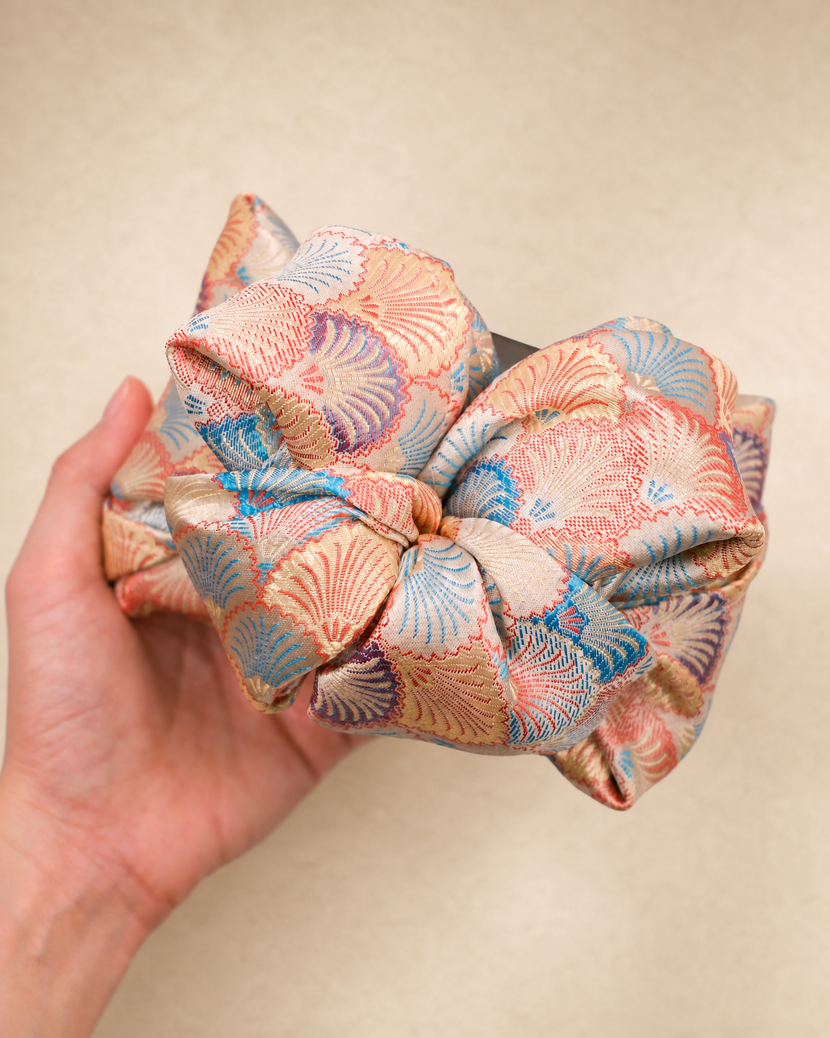 A hand holding a large fabric hair bow with a peach, blue, and purple fan pattern on a beige background.