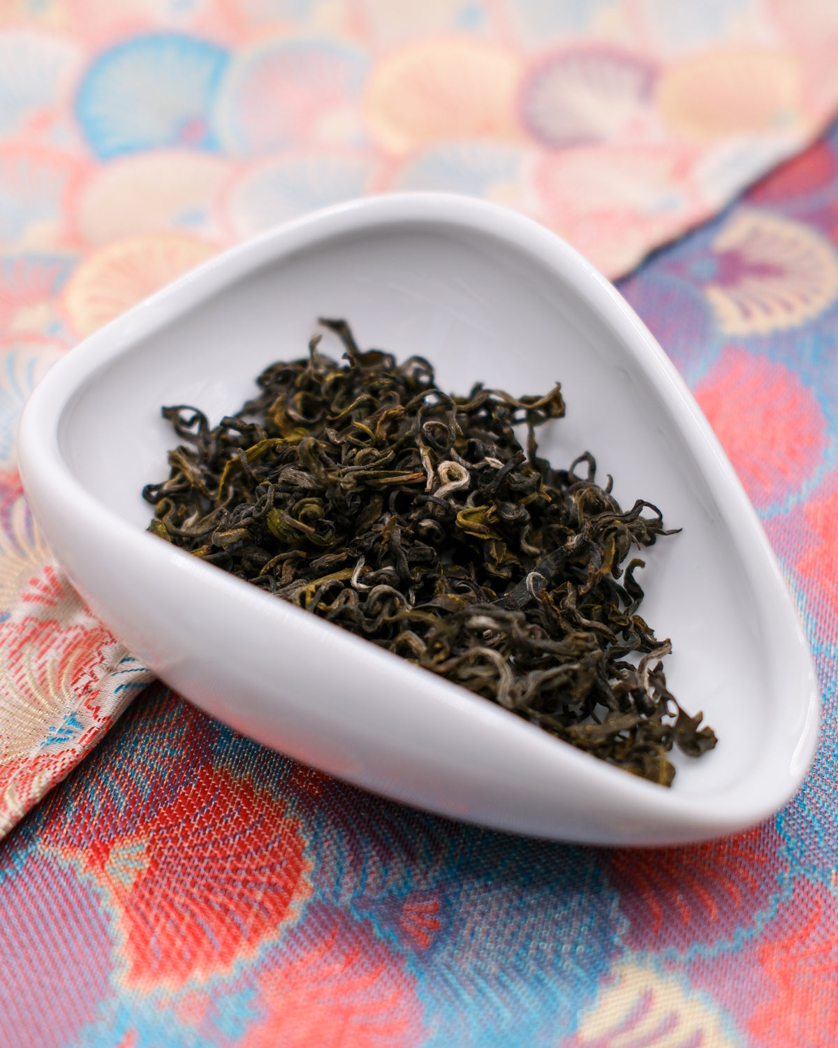 Dried loose-leaf yellow tea leaves from Cha Cha Chan in a small white ceramic dish, set on a colourful patterned fabric background.