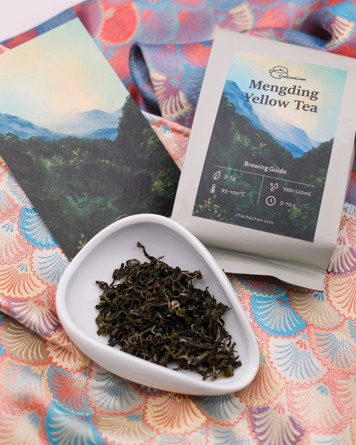 A package of Mengding Yellow Tea from Cha Cha Chan with a brewing guide and a small white dish of loose tea leaves, styled on a colourful patterned fabric background.