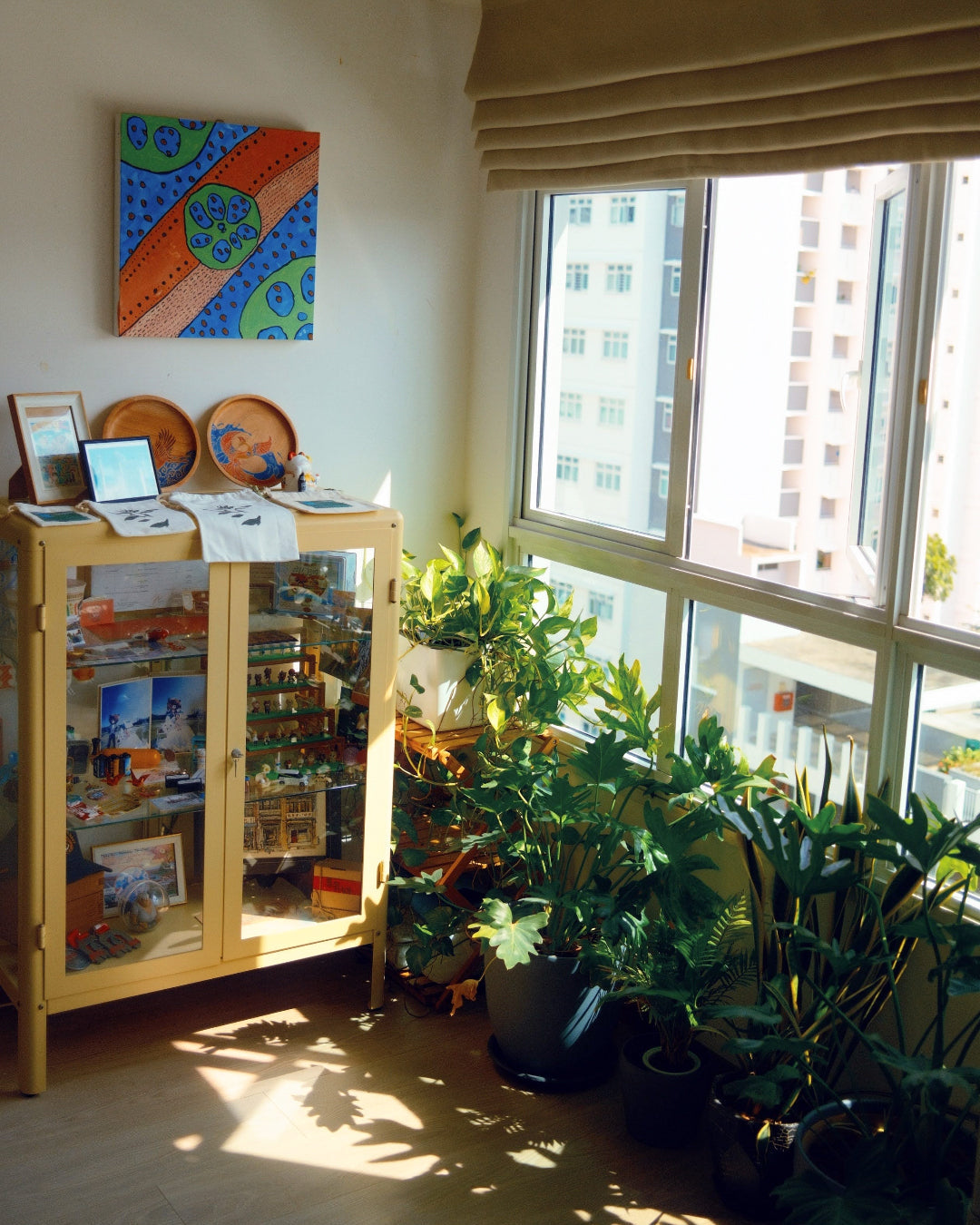 A bright room corner with a wooden cabinet, colorful wall art, and many potted green plants near a large window with a beige roman shade. Sunlight streams in from the right side, casting shadows on the floor.