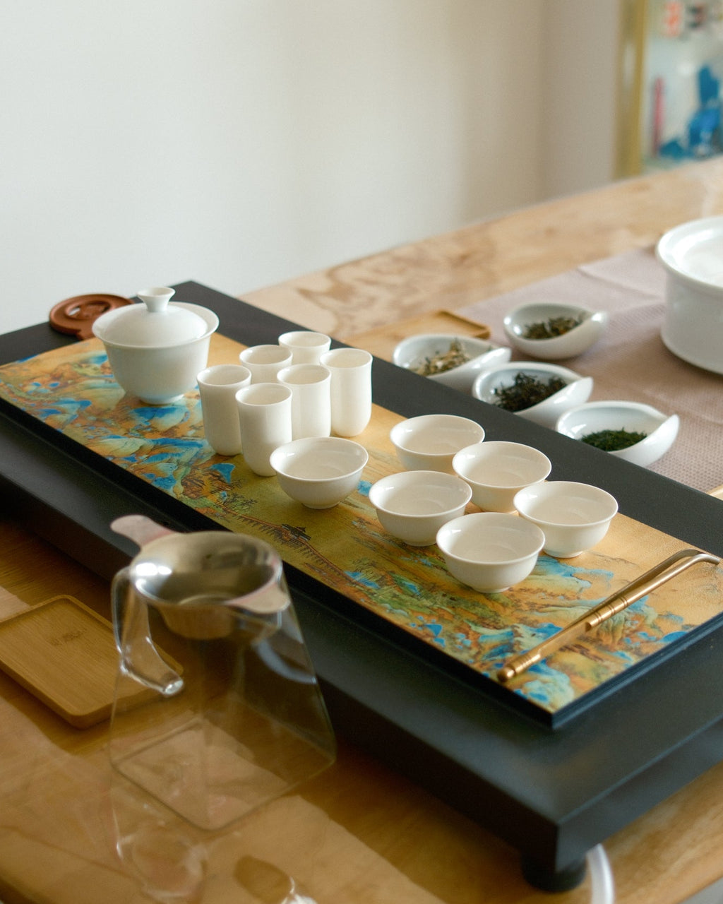 A Japanese-style tea set arranged on a rectangular black tray with a gold-patterned center panel, placed on a light wood dining table in a bright room near a window.