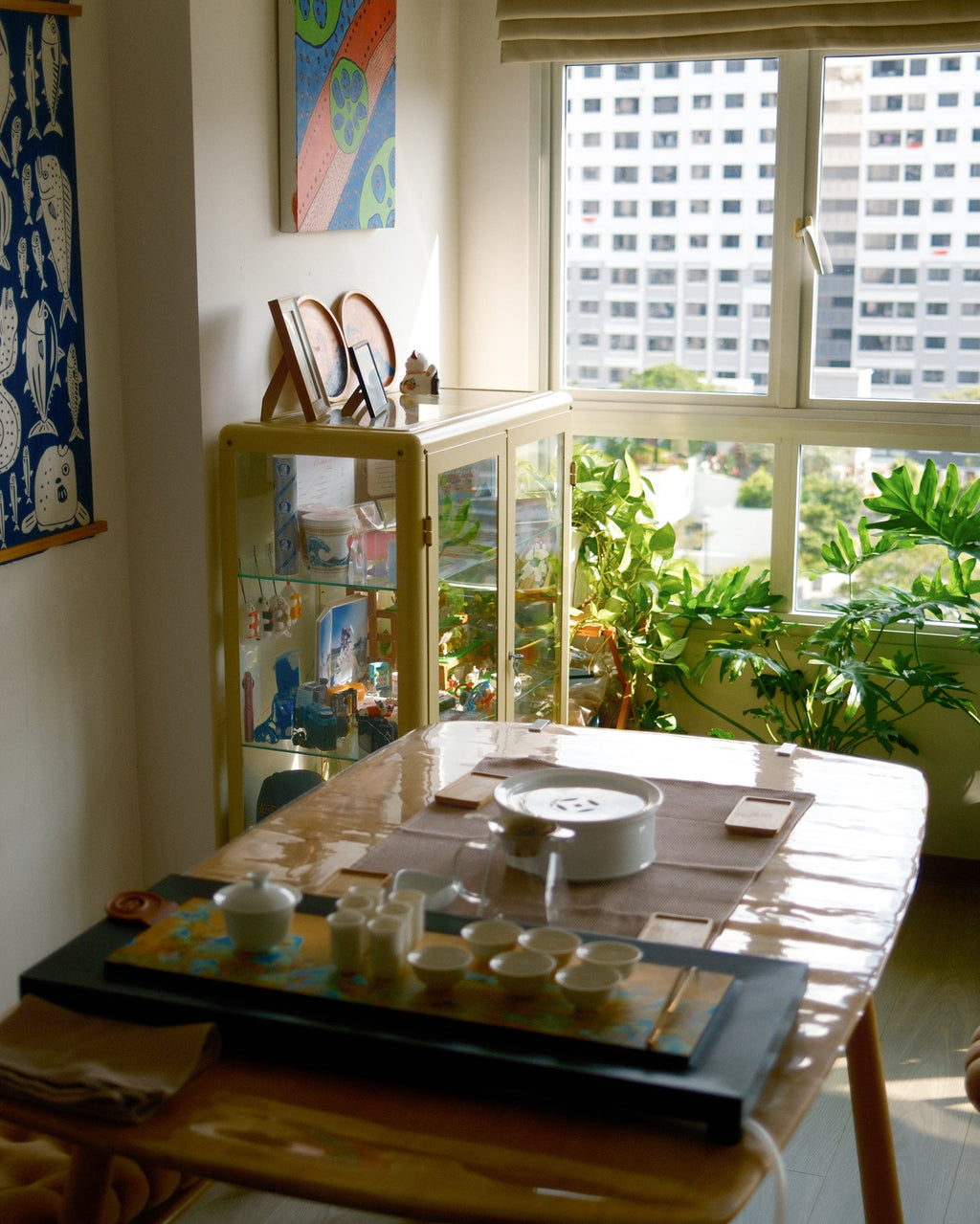 A styled interior room scene with a wooden dining or tea table in the foreground, a glass display cabinet, potted plants by a large window, and wall art hanging on the walls. No person is present, but this is not a clean isolated product shot; it is a real environment, so subject_type is Lifestyle.