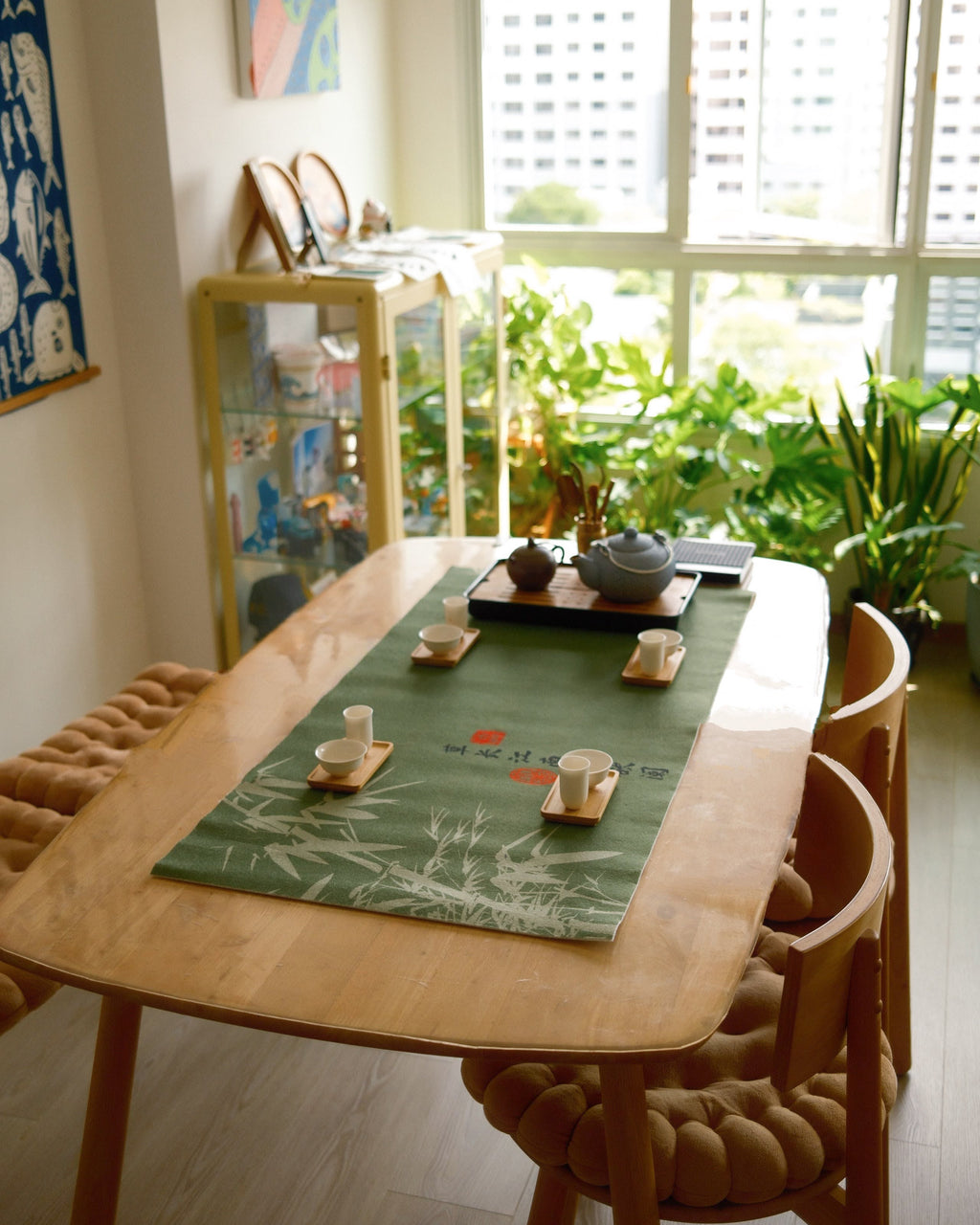 A styled dining room scene featuring a light wood dining table with a green bamboo-patterned runner, set with teacups and a tea set. The table is surrounded by wooden chairs with textured woven seat cushions.