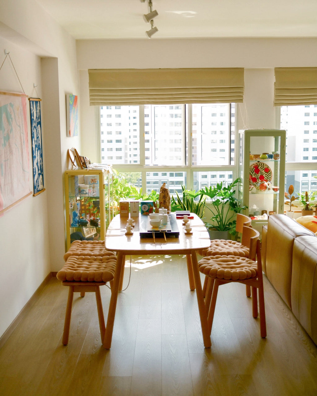 A bright dining area with a light wood table and four woven seat cushions, set in a sunlit apartment with large windows, plants, and a beige sofa.