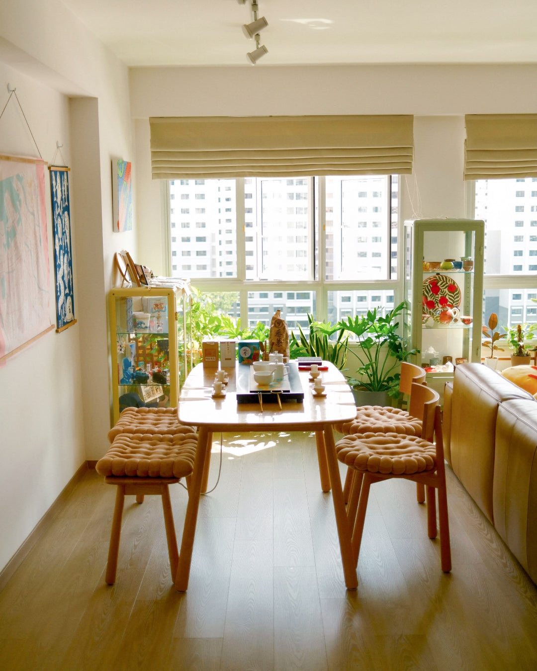 A bright dining area with a light wood table and four woven seat cushions, set in a sunlit apartment with large windows, plants, and a beige sofa.