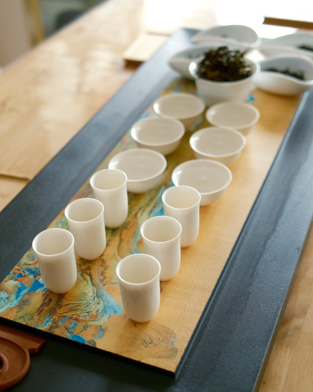 A tea serving set arranged on a long wooden tray over a dark rectangular base, with multiple small white cups and bowls on a light wood table.