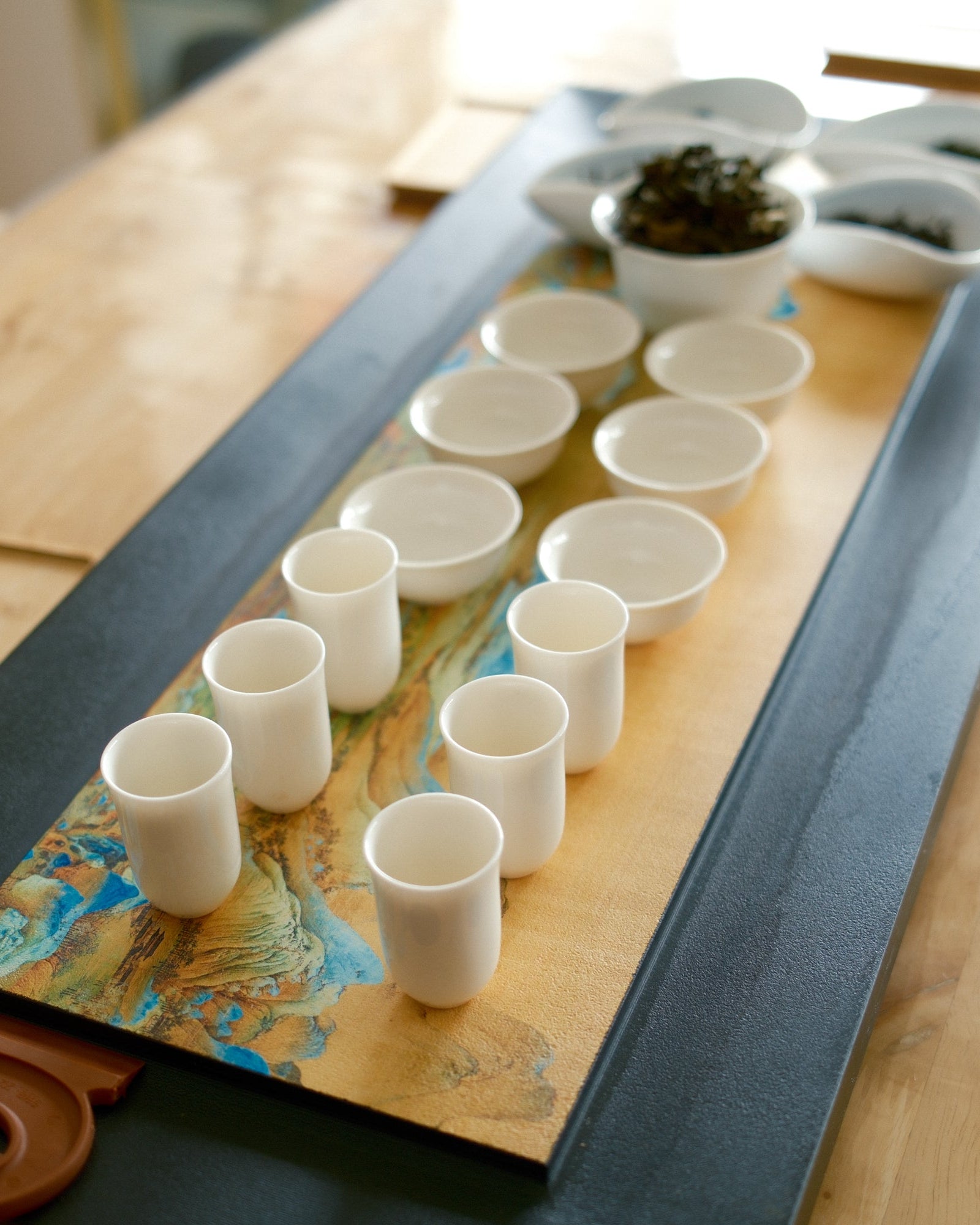 A tea serving set arranged on a long wooden tray over a dark rectangular base, with multiple small white cups and bowls on a light wood table.