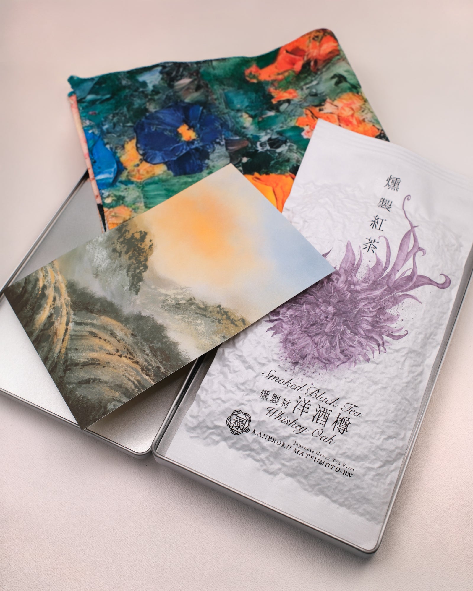 A packaged 'Whisky Oak Smoked Black Tea' loose leaf tea from Cha Cha Chan, featuring a colourful abstract paint-stroke pattern in orange, green, red, and cream, a postcard featuring tea mountains and a metal tin box to store everything.