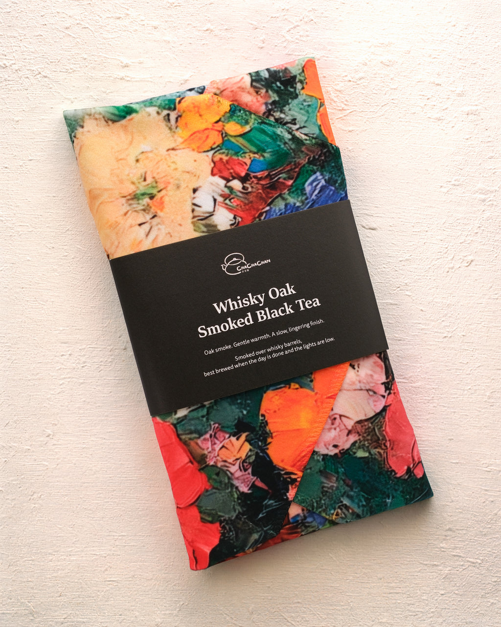 A packaged loose leaf tea from Cha Cha Chan, featuring a colourful abstract paint-stroke pattern in orange, green, red, and cream, wrapped with a black paper band labelled 'Whisky Oak Smoked Black Tea'.