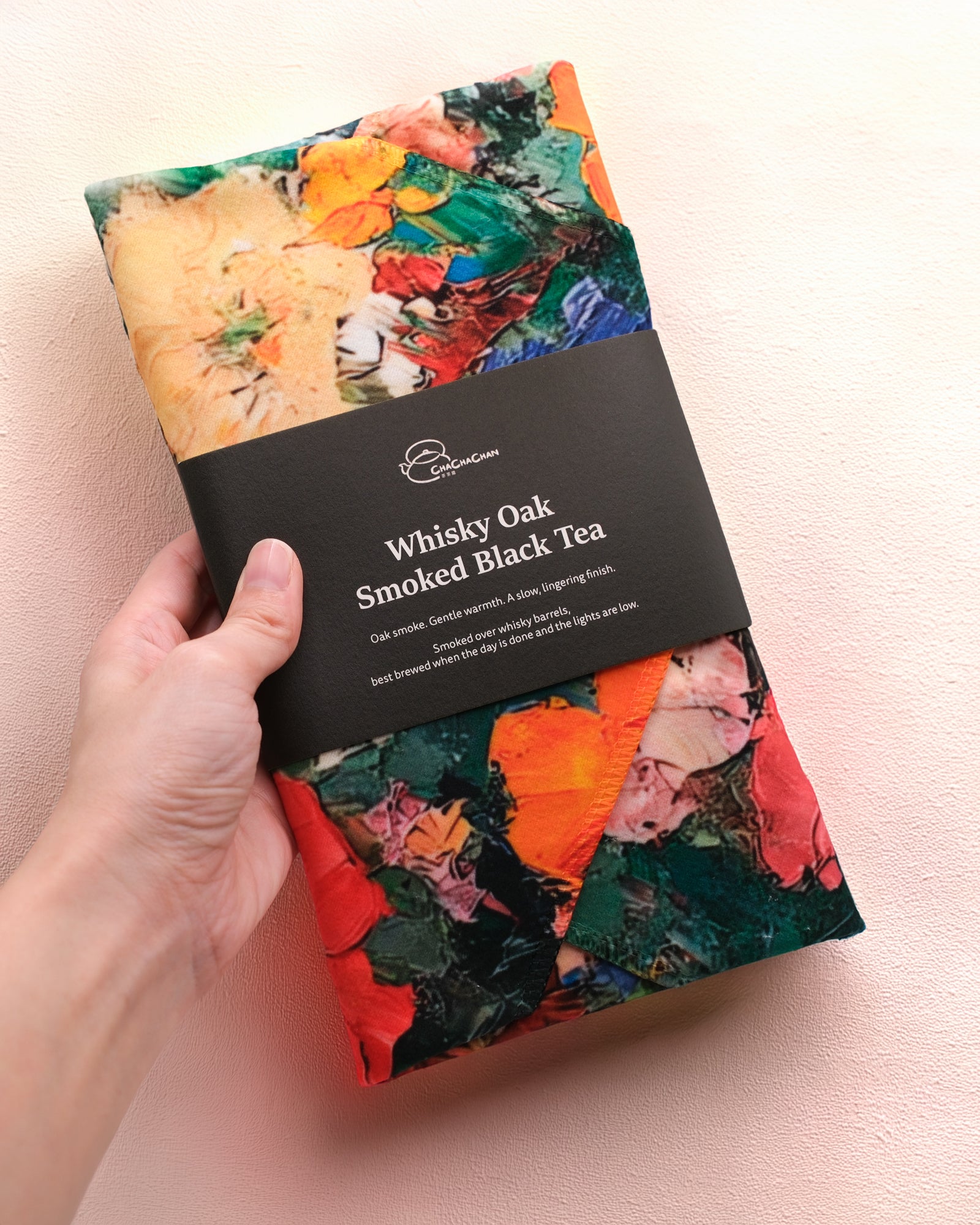 A packaged loose leaf tea from Cha Cha Chan, featuring a colourful abstract paint-stroke pattern in orange, green, red, and cream, wrapped with a black paper band labelled 'Whisky Oak Smoked Black Tea'.