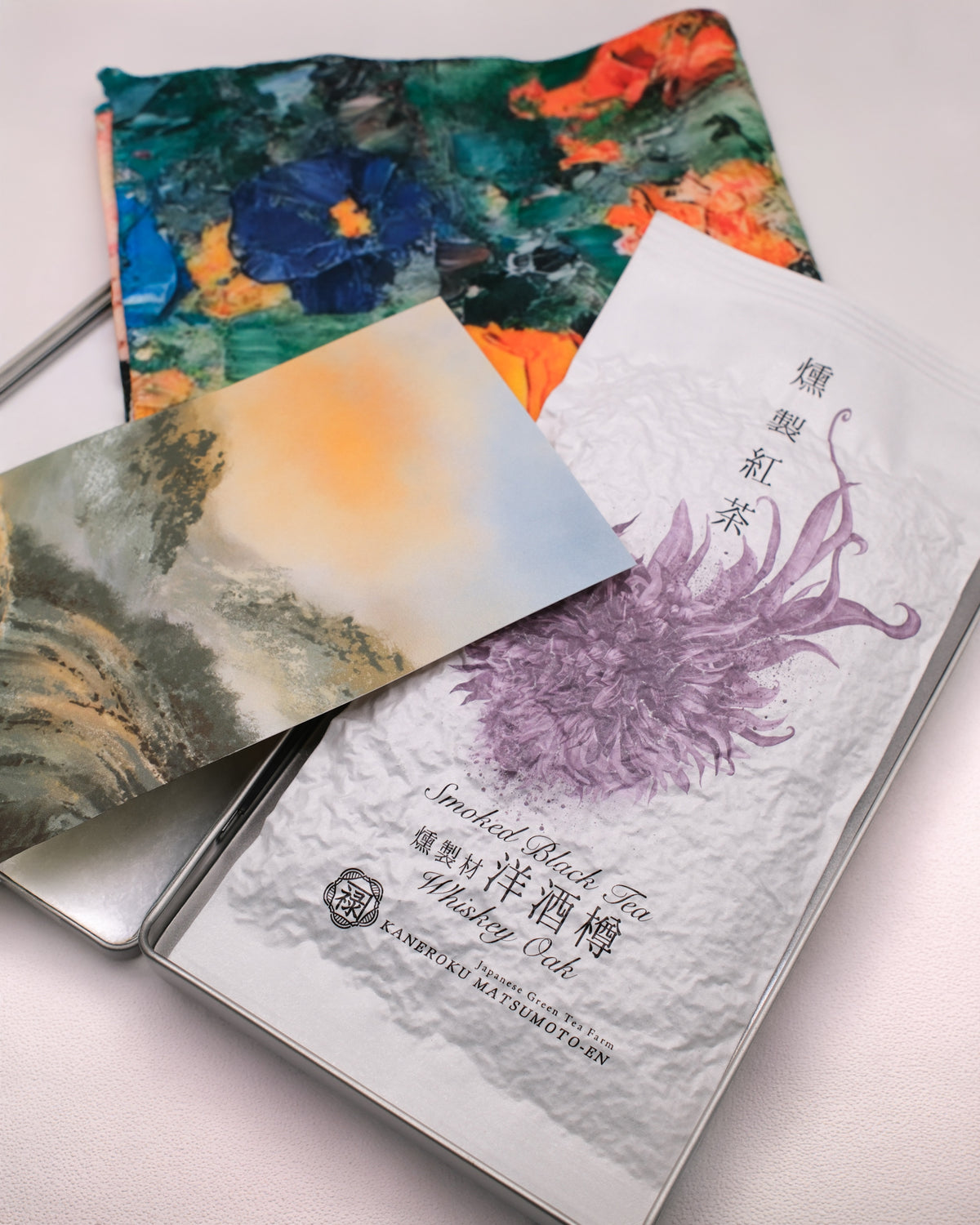 A packaged 'Whisky Oak Smoked Black Tea' loose leaf tea from Cha Cha Chan, featuring a colourful abstract paint-stroke pattern in orange, green, red, and cream, a postcard featuring tea mountains and a metal tin box to store everything.