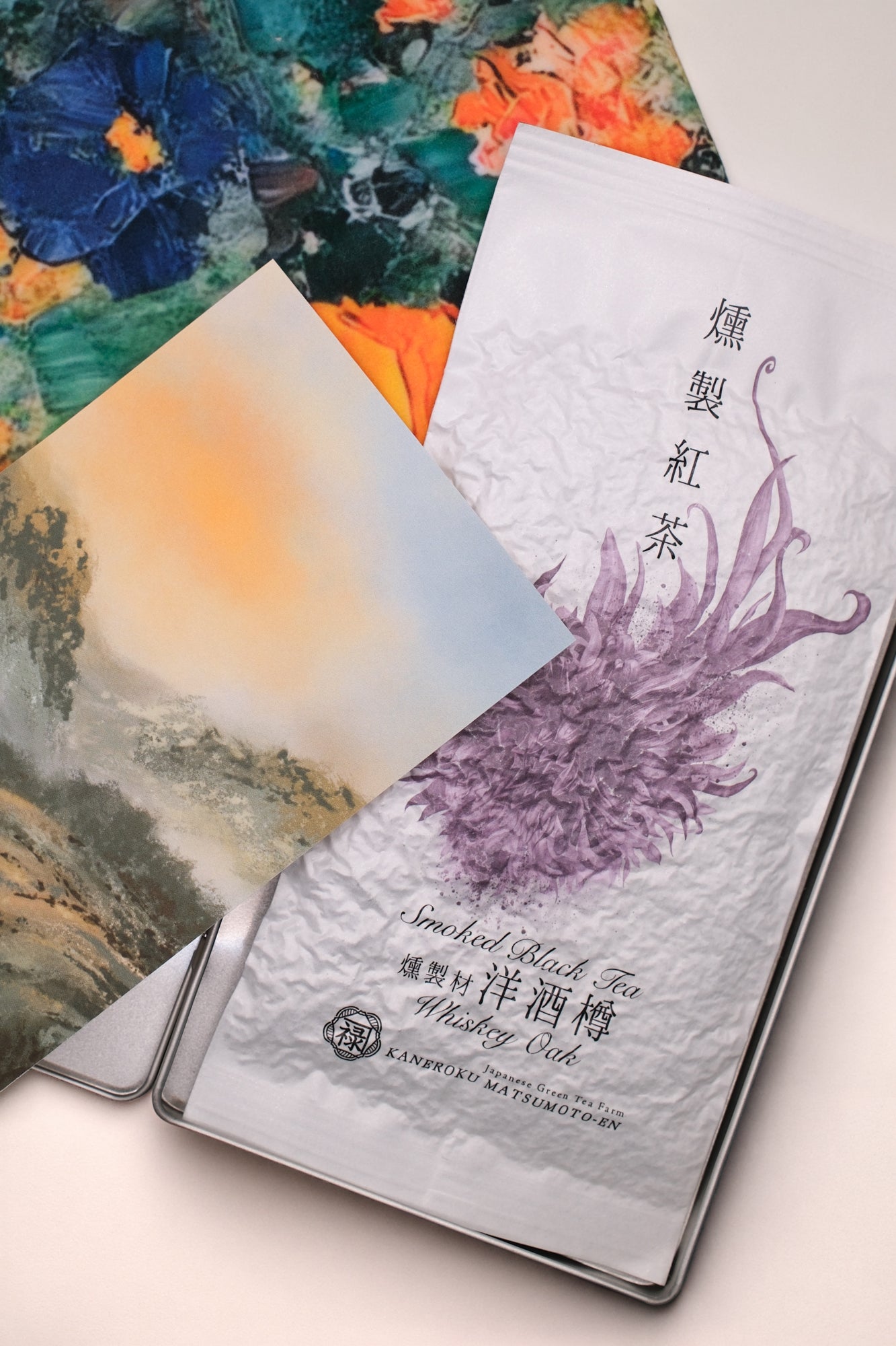 A packaged 'Whisky Oak Smoked Black Tea' loose leaf tea from Cha Cha Chan, featuring a colourful abstract paint-stroke pattern in orange, green, red, and cream, a postcard featuring tea mountains and a metal tin box to store everything.