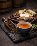 Tea cup with a wooden scoop of tea leaves, cheese platter with crackers, and barrel in the background