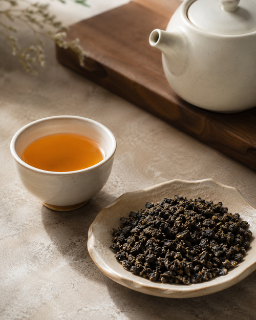 A ceramic cup filled with amber White Bud Rare Orchid Oolong Tea sits beside a shallow dish of dark loose-leaf tea, with a white teapot on a wooden board in the background.