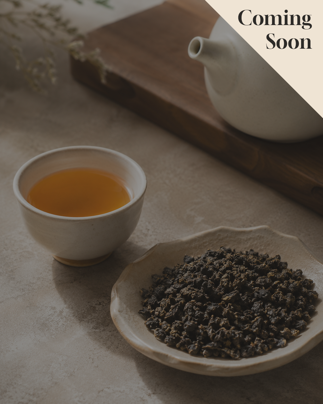 A styled tea scene with a small white teacup filled with amber White Bud Rare Orchid Oolong Tea, a plate of dark loose-leaf tea, and part of a white teapot on a wooden board in the background. A beige diagonal banner in the top right reads 'Coming Soon'.